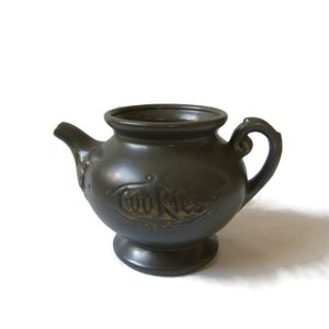 May include: A dark brown ceramic cookie jar shaped like a teapot with a gold inscription that reads "Cookies".