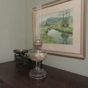 May include: A clear glass oil lamp with a decorative etched base and a tall, cylindrical chimney. The lamp sits on a dark wooden surface, with a vintage scale and a framed landscape painting in the background. The lamp is a vintage item.