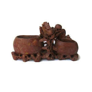 May include: A reddish-brown, carved stone double bowl with intricate floral and fruit details. The bowls are connected by a carved branch and sit on a detailed base. The piece is likely a decorative item.