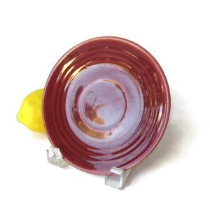 May include: A round, glossy ceramic saucer featuring a deep red glaze with subtle iridescent purple and gold highlights. The saucer has concentric circular ridges along its surface and is displayed on a clear acrylic stand next to a yellow lemon.