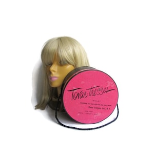 May include: A blonde wig on a mannequin head next to a round pink wig box. The box has the words "Tovar Tresses" in black script. The box also has a black handle. The wig has bangs and is shoulder length.