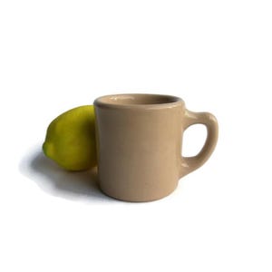 May include: A beige ceramic mug with a rounded handle sits next to a bright yellow lemon. The mug has a simple, classic design, perfect for coffee or tea. The lemon adds a pop of color.