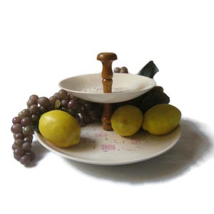 May include: A two-tiered serving stand featuring two off-white plates with a subtle pink and black geometric pattern, connected by a central brown wooden spindle. The stand is decorated with a cluster of purple grapes and two yellow lemons.