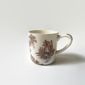 May include: A white ceramic mug with a brown and green floral pattern. The mug features a scene of a small cottage with a water wheel and a river.