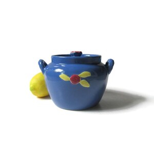 May include: A blue ceramic jar with a lid and two handles. The jar is decorated with a red and yellow floral design. A yellow lemon is next to the jar. The jar is approximately 5 inches in diameter.