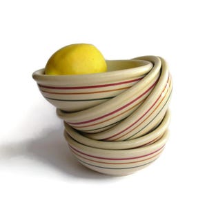 May include: A stack of four cream-colored ceramic bowls with thin horizontal stripes in red, orange, and green. A bright yellow lemon rests inside the top bowl. The bowls have a smooth, rounded design and are arranged in a slightly tilted vertical stack.