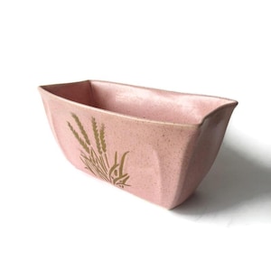 May include: A pink rectangular planter with a speckled finish and a gold wheat stalk design. The planter has a slightly curved shape and is made of ceramic. Ideal for displaying plants or flowers.