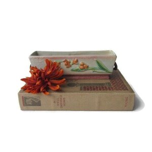 May include: A rectangular, off-white ceramic planter with a floral design, resting on a vintage book titled "A Well Dressed Home." An orange-red flower is placed in front of the planter, adding a pop of color to the composition.