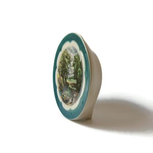 May include: A small ceramic trinket box with a teal blue rim and a white background. The box features a painted scene of a country road with trees and a river.
