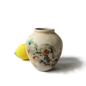 May include: A small, cream-colored ceramic vase with a marbled design in shades of green, brown, and orange. The vase is round and has a wide opening.