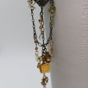 Può includere: Una collana con pendente in stile antico con una parte superiore a filigrana a forma di diamante. La collana presenta un ciondolo a forma di chiave, una rosa e varie perline e ciondoli nei toni dell'oro, del marrone e della crema. La catena è di colore bronzo scuro.