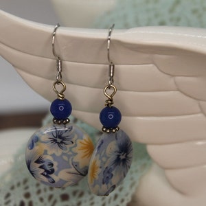 May include: Dangle earrings with floral ceramic beads in blue, yellow, and white. Each earring has a dark blue bead and a silver hook. The earrings are displayed against a white background. The earrings are a unique accessory.