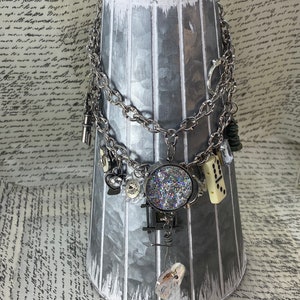 May include: A silver-toned necklace with multiple charms, including a round pendant filled with glitter, a domino, and a small clear crystal. The necklace hangs on a silver metal vase with white accents.