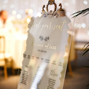 Puede incluir: Un cartel de asignación de asientos de boda de acrílico transparente con letras doradas. El cartel está titulado "find your seat" y enumera los nombres de los invitados bajo los encabezados "one", "two", "three", "four", "five" y "six".