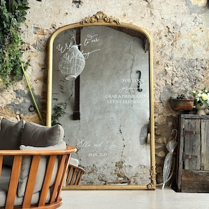 Puede incluir: Un gran espejo dorado adornado con texto escrito para una boda. El espejo refleja una habitaci&oacute;n con una silla de madera, plantas y una pared r&uacute;stica. El texto incluye frases como "Bienvenido a nuestra boda" y "Toma una copa y celebremos."