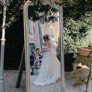 May include: A large, ornate mirror with a silver frame, reflecting a bride in a wedding dress. The mirror has a decorative top and displays wedding details. The scene is set outdoors with greenery and a stone pathway.