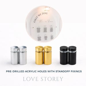 May include: A clear acrylic wedding seating chart with the text "FIRST WE DINE" and guest names. Below are silver, gold, and black standoff fixings for mounting. The text "PRE-DRILLED ACRYLIC HOLES WITH STANDOFF FIXINGS" and "LOVE STOREY" are also visible.