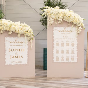 May include: Two white wedding welcome signs with gold lettering on a blush pink background. The signs are decorated with white flowers. The left sign says "Welcome to our wedding Sophie & James 21st May 2022". The right sign says "Welcome Sophie & James by Table".