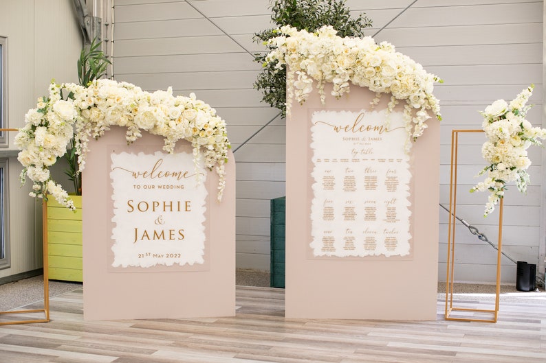 May include: Two white wedding welcome signs with gold lettering on a blush pink background. The signs are decorated with white flowers. The left sign says "Welcome to our wedding Sophie & James 21st May 2022". The right sign says "Welcome Sophie & James by Table".