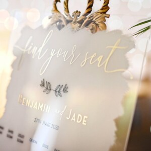 May include: A white acrylic sign with gold lettering that reads "find your seat" and "Benjamin and Jade" with the date "27th June 2021".