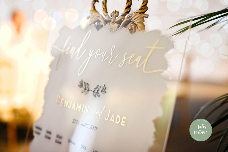 May include: A white acrylic sign with gold lettering that reads "find your seat" and "Benjamin and Jade" with the date "27th June 2021".