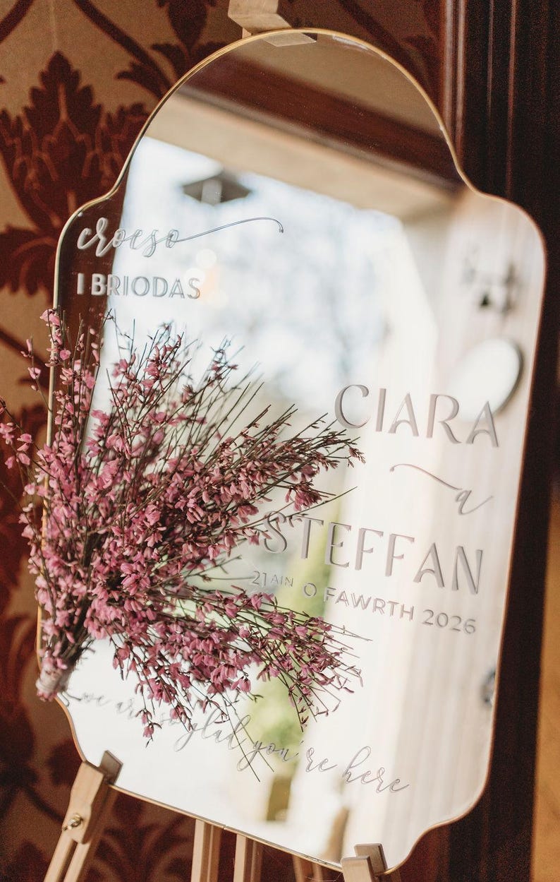 Puede incluir: Espejo decorativo con marco dorado, grabado con nombres y fecha. Muestra los nombres "Ciara & Steffan" y la fecha "21ain o Fawrth 2026". Un ramo de flores rosas adorna el espejo. El texto "Croeso I Briodas" est&aacute; grabado.