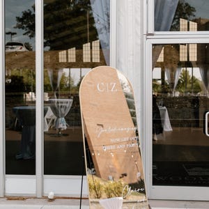 Puede incluir: Un gran espejo arqueado con marco de madera se encuentra fuera de un edificio. El espejo refleja el entorno y tiene las iniciales "CIZ" en la parte superior. El texto en el espejo dice "You look amazing, now get out here and party" y los nombres "Claire y Zachary".