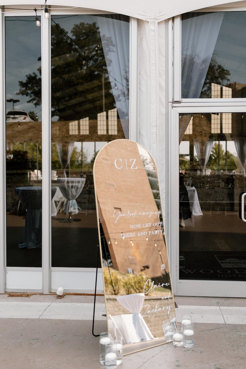 Puede incluir: Un gran espejo arqueado con marco de madera se encuentra fuera de un edificio. El espejo refleja el entorno y tiene las iniciales "CIZ" en la parte superior. El texto en el espejo dice "You look amazing, now get out here and party" y los nombres "Claire y Zachary".