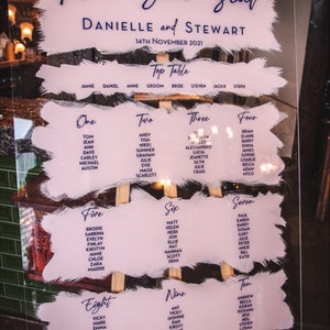 May include: A clear acrylic wedding seating chart with a pink and white painted background. The chart is titled "Take Your Seat" and lists the names of guests for each table. The chart is displayed on a wooden easel.