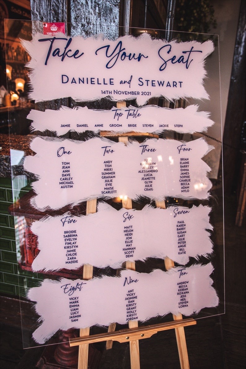 May include: A clear acrylic wedding seating chart with a pink and white painted background. The chart is titled "Take Your Seat" and lists the names of guests for each table. The chart is displayed on a wooden easel.