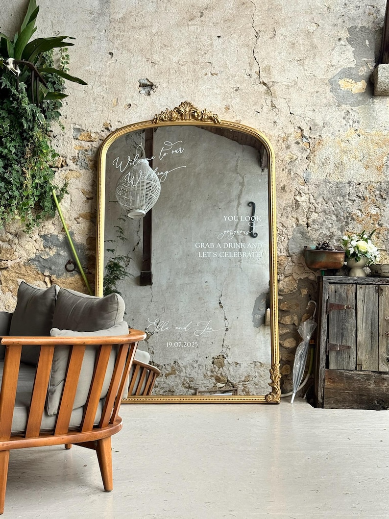 Puede incluir: Un gran espejo dorado adornado con texto escrito para una boda. El espejo refleja una habitaci&oacute;n con una silla de madera, plantas y una pared r&uacute;stica. El texto incluye frases como "Bienvenido a nuestra boda" y "Toma una copa y celebremos."