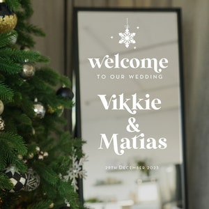 May include: A white sign with a snowflake design and the text "welcome to our wedding Vikkie & Matias 29th December 2023" reflected in a mirror.