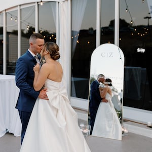 Puede incluir: Una pareja se abraza y se besa frente a un gran espejo en una boda al aire libre. La novia lleva un vestido blanco sin tirantes con un gran lazo. El novio lleva un traje azul. El espejo refleja a la pareja y tiene las iniciales "CIZ".