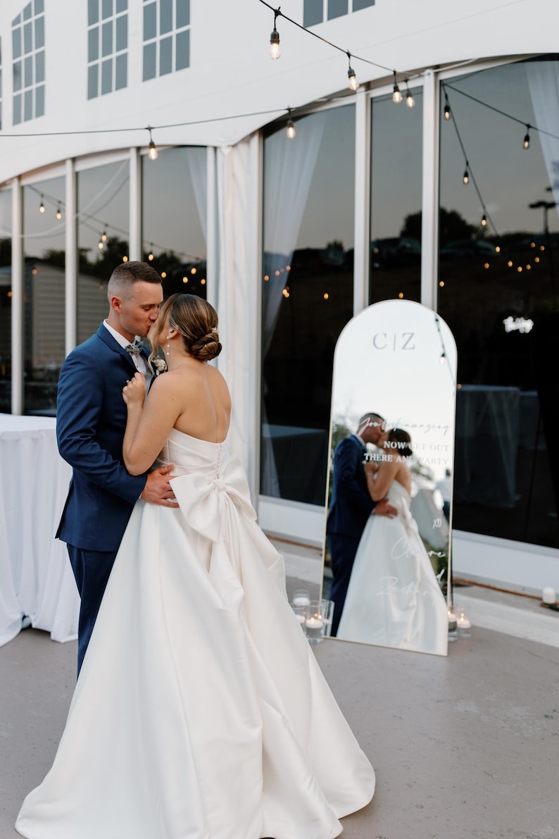 Puede incluir: Una pareja se abraza y se besa frente a un gran espejo en una boda al aire libre. La novia lleva un vestido blanco sin tirantes con un gran lazo. El novio lleva un traje azul. El espejo refleja a la pareja y tiene las iniciales "CIZ".