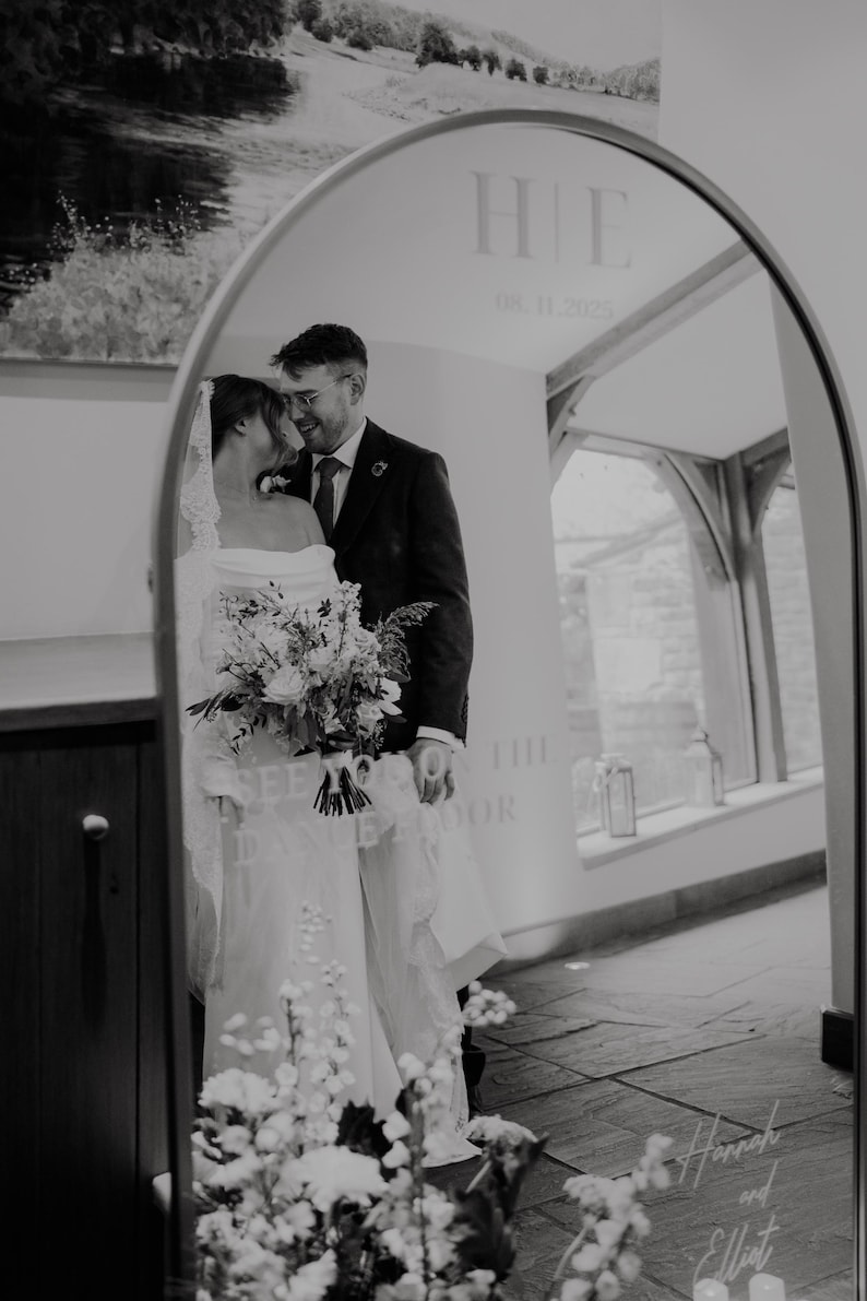 A black and white image of a bride and groom standing in front of a mirror, with the bride holding a bouquet of flowers.