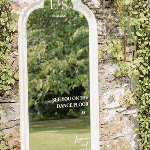 Puede incluir: Espejo enmarcado en blanco con una parte superior arqueada, con el texto "SEE YOU ON THE DANCE FLOOR" y los nombres "Lauren and Scott". El espejo est&aacute; colocado contra una pared de piedra con vegetaci&oacute;n.