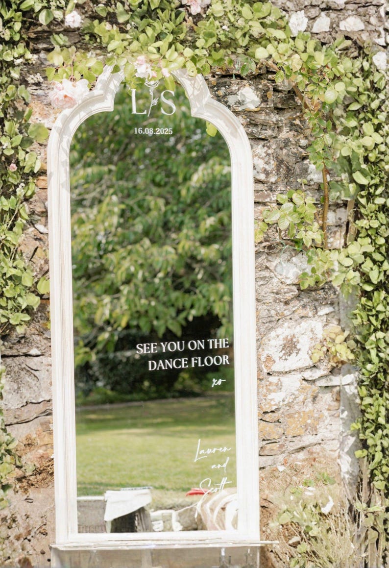 Puede incluir: Espejo enmarcado en blanco con una parte superior arqueada, con el texto "SEE YOU ON THE DANCE FLOOR" y los nombres "Lauren and Scott". El espejo est&aacute; colocado contra una pared de piedra con vegetaci&oacute;n.