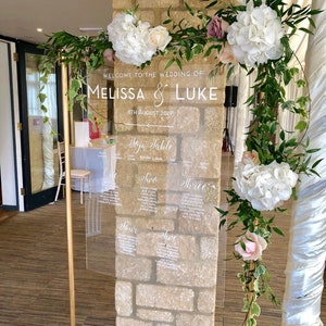 May include: A clear acrylic wedding seating chart with white calligraphy lettering. The chart is mounted on a gold stand and decorated with white and pink flowers and greenery.