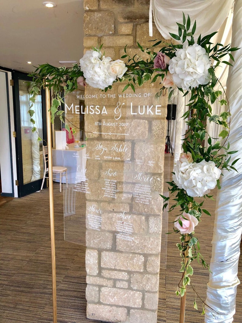 May include: A clear acrylic wedding seating chart with white calligraphy lettering. The chart is mounted on a gold stand and decorated with white and pink flowers and greenery.
