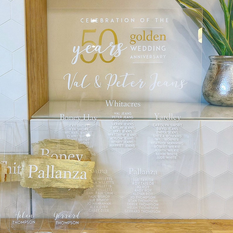 May include: A clear acrylic sign with gold lettering announcing a 50th wedding anniversary celebration for Val & Peter Jeans. The sign lists the names of guests seated at tables named Whitacres, Boney Hay, Pallanza, and Yardley.