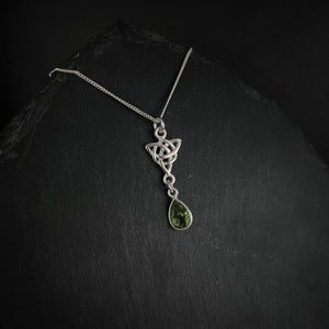 Pode incluir: Um colar de prata com um pingente de nó celta e uma gota de pedra preciosa verde. O pingente tem a forma de um triângulo com um design de nó. A pedra preciosa é em forma de pêra.