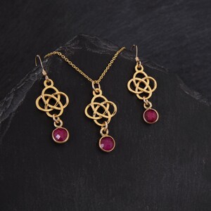 May include: A gold necklace and earrings set with a Celtic knot design and red gemstone accents.
