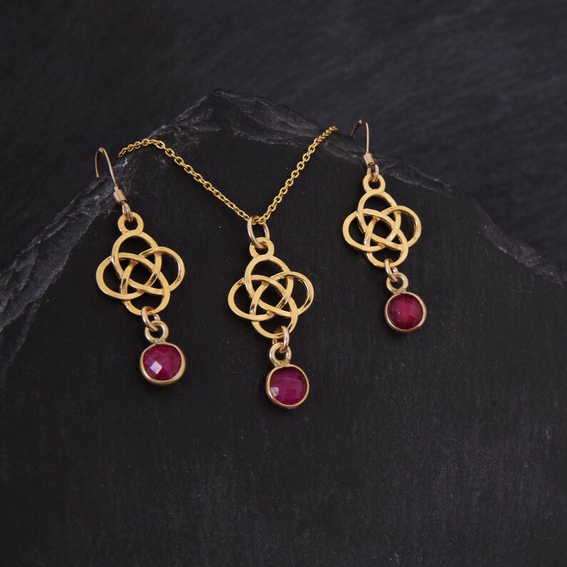 May include: A gold necklace and earrings set with a Celtic knot design and red gemstone accents.