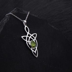 Puede incluir: Un collar de plata con un colgante de nudo celta con una piedra preciosa verde. El colgante tiene forma de nudo con una sola piedra preciosa verde en el centro.