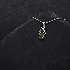 May include: A silver pendant necklace with a green oval gemstone set in a Celtic knot design. The necklace is on a black background.