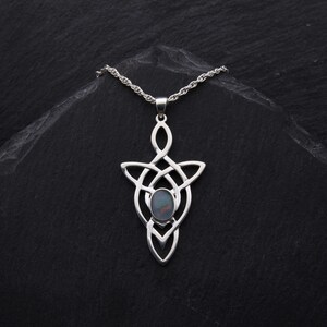 Puede incluir: Un collar de plata con un colgante de nudo celta con una piedra de &oacute;palo iridiscente oscura en el centro. El collar est&aacute; sobre un fondo negro.