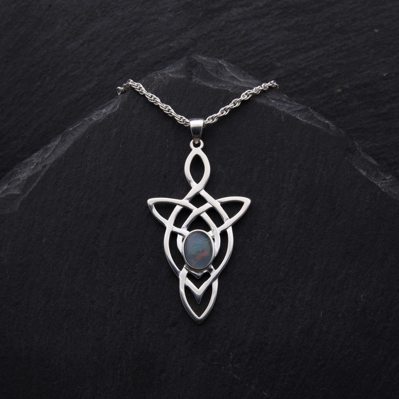 Puede incluir: Un collar de plata con un colgante de nudo celta con una piedra de &oacute;palo iridiscente oscura en el centro. El collar est&aacute; sobre un fondo negro.