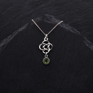 Op de afbeelding: Een zilverkleurige hangerketting met een Keltisch knoopontwerp en een kleine, ronde, groene edelsteen. De ketting hangt aan een delicate ketting tegen een donkere, getextureerde achtergrond. De hanger is ongeveer 2,5 cm lang.