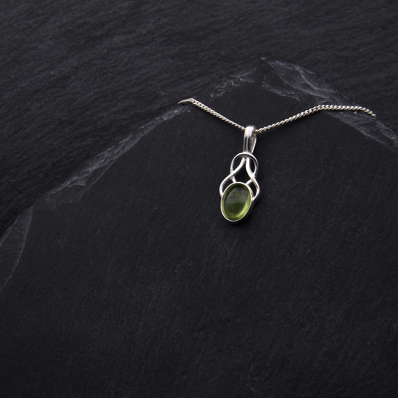 May include: A silver pendant necklace with a green oval gemstone set in a Celtic knot design. The necklace is displayed on a black slate background.