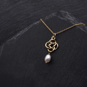 May include: A gold necklace with a Celtic knot pendant and a single pearl drop. The necklace is on a black background.